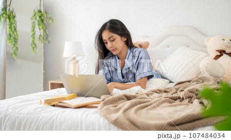 A beautiful Asian woman in pajamas is focusing on her work on her laptop while lying on her bed. A beautiful Asian woman in pajamas is focusing on her work on her laptop while lying on her bed. 107484034