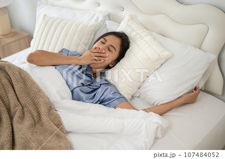 A sleepy and carefree Asian woman is yawning after waking up in the morning on her comfy bed. A sleepy and carefree Asian woman is yawning after waking up in the morning on her comfy bed. 107484052