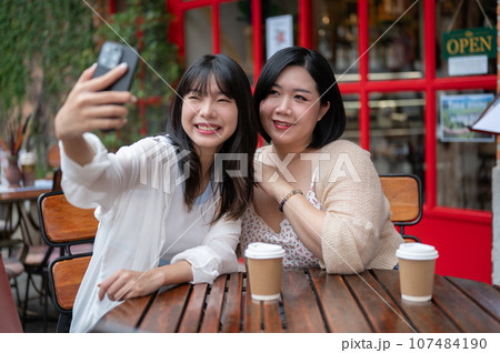 Two happy Asian female friends using a smartphone to take selfies while relaxing at a coffee shop. Two happy Asian female friends using a smartphone to take selfies while relaxing at a coffee shop. 107484190
