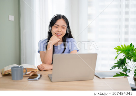 An attractive Asian woman wearing headphones studying an online class through her laptop. 107484343