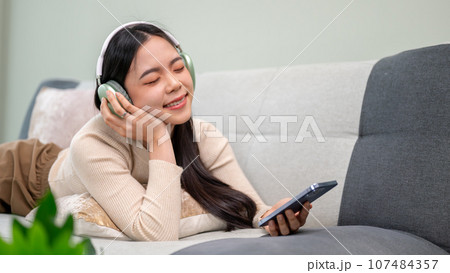 A relaxed and beautiful Asian woman wearing headphones and enjoying the music while lying on a sofa. 107484357