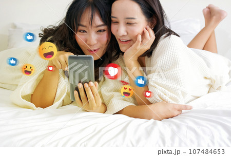 Two cheerful asian women, nestled in bed in bathrobes, are indulged in gleeful selfie-session, surrounded by vibrant digital emoticons. Two cheerful asian women, nestled in bed in bathrobes, are indulged in gleeful selfie-session, surrounded by vibrant digital emoticons. 107484653