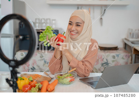 A beautiful Asian Muslim female food blogger is live streaming on her social media in the kitchen. 107485280