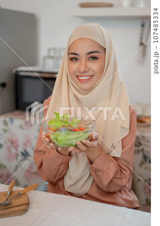 A beautiful Asian Muslim holding a bowl of healthy yummy salad while sitting at a dining table A beautiful Asian Muslim holding a bowl of healthy yummy salad while sitting at a dining table 107485334