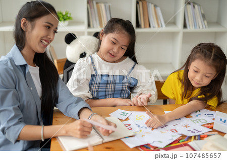 Two adorable young Asian girls are enjoying studying English with a teacher at home. Two adorable young Asian girls are enjoying studying English with a teacher at home. 107485657