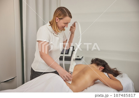 vacuum-roller manipulator, beauty clinic, anti cellulite. Attractive young woman is in process of improving body shape, using vacuum roller to massage client, correcting figure, anti-cellulite massage vacuum-roller manipulator, beauty clinic, anti cellulite. Attractive young woman is in process of improving body shape, using vacuum roller to massage client, correcting figure, anti-cellulite massage 107485817