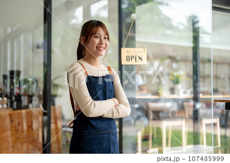 A pretty Asian female coffee shop owner standing with her arms crossed at the entrance door. 107485999