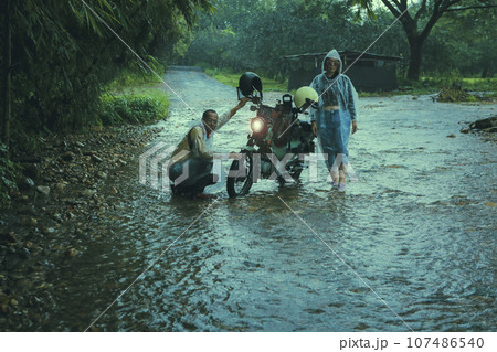 couples of asian biker wearing plastic rain clothes  maintenance small enduro motorcycle in shallow creek among rain falling 107486540