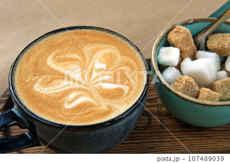 Blue Ceramic Cup of Hot Cappuccino with Flower Shaped Foam on Wooden Cafe Table. Beautiful Latte Art Froth. Trendy Authentic Modern Porcelain Set. Pottery Handmade Coffee Mug Served with a Sugar Bowl Blue Ceramic Cup of Hot Cappuccino with Flower Shaped Foam on Wooden Cafe Table. Beautiful Latte Art Froth. Trendy Authentic Modern Porcelain Set. Pottery Handmade Coffee Mug Served with a Sugar Bowl 107489039