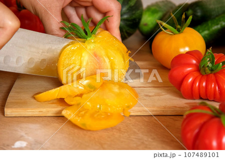 Woman using a knife cutting fresh raw vegetables. Tomatoes, cucumbers being sliced on a wooden board. Cooker at home making a healthy veggie meal. Chopping ingredients for preparing vegetarian dish Woman using a knife cutting fresh raw vegetables. Tomatoes, cucumbers being sliced on a wooden board. Cooker at home making a healthy veggie meal. Chopping ingredients for preparing vegetarian dish 107489101