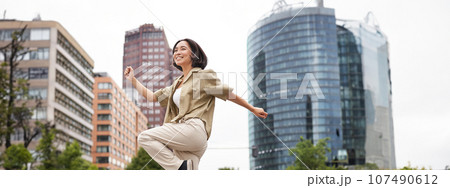 Vertical shot of young asian woman posing happy, raising hands up and dancing, triumphing, celebrating victory, enjoying day out in city Vertical shot of young asian woman posing happy, raising hands up and dancing, triumphing, celebrating victory, enjoying day out in city 107490612