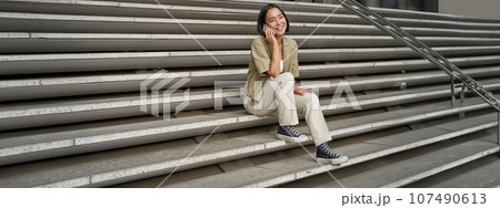 Portrait of asian student girl on stairs talks on mobile phone, smiles at camera, sits outside building 107490613