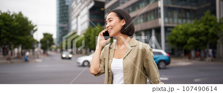 Portrait of smiling asian woman talking on mobile phone, walking on street near busy road and speaking to friend, laughing 107490614