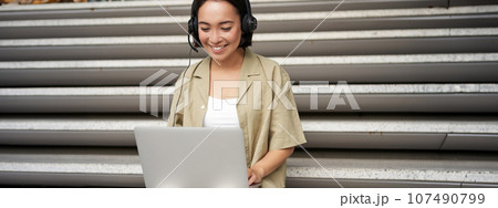 Portrait of asian woman, digital nomad girl using laptop and listens to music outdoors. Young student works on computer and smiles 107490799