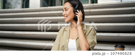 Close up portrait of smiling asian woman listens music in headphones, turns around with happy face expression, resting outdoors 107490828