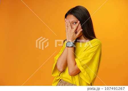 Teen girl over yellow background in studio covering face with hands 107491267