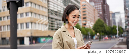 Young asian woman exploring city with smartphone app, holding mobile phone and walking on screet 107492067