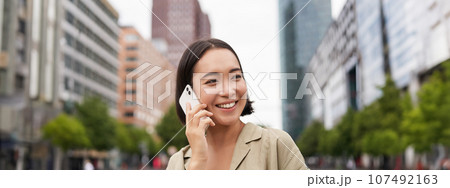 Portrait of smiling asian woman talking on mobile phone, walking on street near busy road and speaking to friend, laughing 107492163