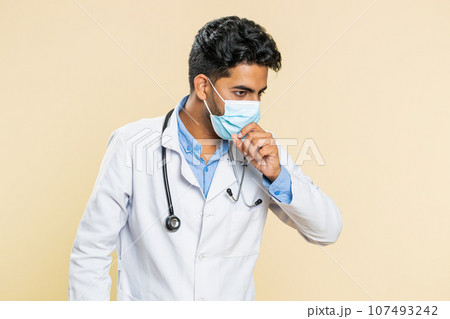 Indian young doctor man in protective mask cough with seasonal flu symptoms fever allergy infection Indian young doctor man in protective mask cough with seasonal flu symptoms fever allergy infection 107493242