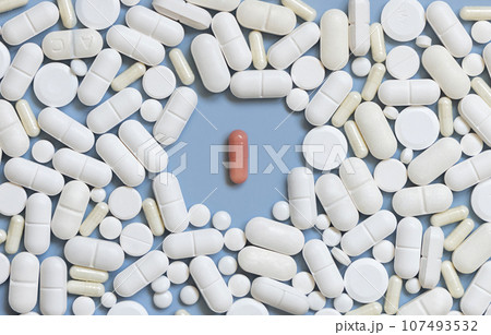 Mix of white medical capsules and pills with one pink capsule in the middle on light blue top view 107493532