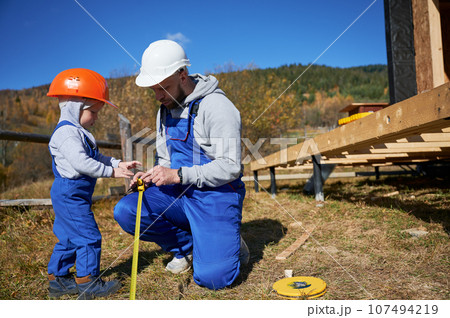 Father with toddler son building wooden frame house on pile foundation. Male builder and kid playing with tape measure on construction site on sunny day. Carpentry and family concept. 107494219