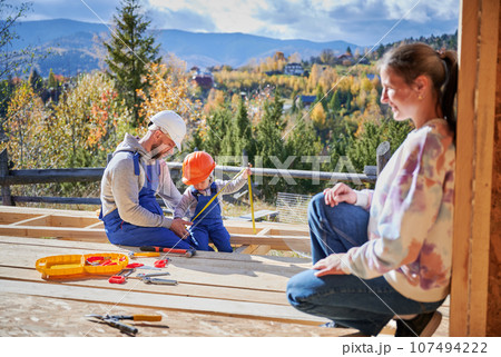 Father, mother and son building wooden frame house. Toddler boy helping his daddy, while woman looking for them on construction site. Guys wearing helmet and blue overalls. Carpentry, family concept. 107494222