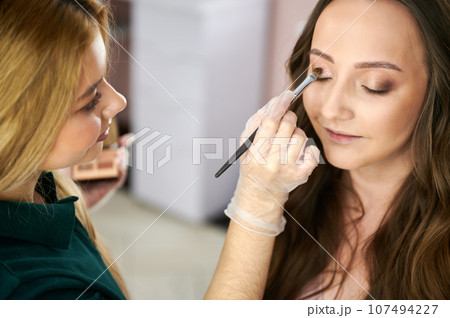 Makeup artist applying eyeshadow on client eyelid with cosmetic brush. Close up of young woman keeping eyes closed and smiling while stylist doing professional eye makeup. Makeup artist applying eyeshadow on client eyelid with cosmetic brush. Close up of young woman keeping eyes closed and smiling while stylist doing professional eye makeup. 107494227