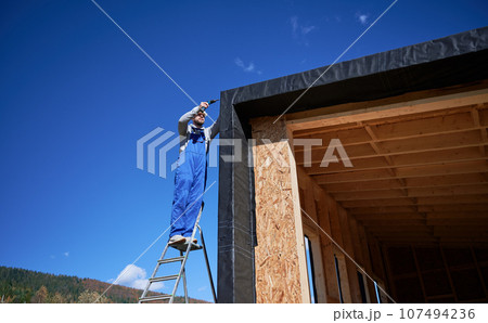 Male builder installing black corrugated iron sheet used as facade of future cottage, blue sky on background. Man worker building wooden frame house. Carpentry and construction concept. Male builder installing black corrugated iron sheet used as facade of future cottage, blue sky on background. Man worker building wooden frame house. Carpentry and construction concept. 107494236