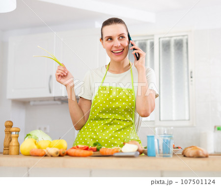Cheerful young housewife talking on phone while cooking in home kitchen Cheerful young housewife talking on phone while cooking in home kitchen 107501124