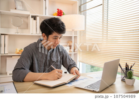 Studying online using a laptop, Man watching live video or video call of teacher teaching on laptop in her home, Take notes of important conversations and messages during the teacher's teaching. Studying online using a laptop, Man watching live video or video call of teacher teaching on laptop in her home, Take notes of important conversations and messages during the teacher's teaching. 107504989