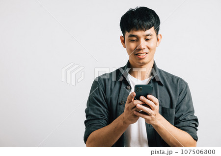 Portrait of a smiling Asian young man, excited while typing a text message on his smart mobile phone for online communication. Studio shot isolated on white, showcasing his enthusiasm for technology. 107506800