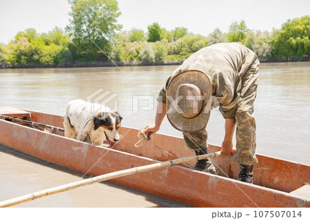 Puzzled fisherman in checkered hat choosing fishing tackle, while leaning forward in boat. Front view of male angler with dog, looking for something at bottom of old red skiff. Concept of fishing. Puzzled fisherman in checkered hat choosing fishing tackle, while leaning forward in boat. Front view of male angler with dog, looking for something at bottom of old red skiff. Concept of fishing. 107507014