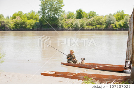 Focused fisherman in hat looking on float, during fishery with dog in cloudy water at pier. Front view of calm male angling, while having rest, with thickets coast on background. Concept of hobby. Focused fisherman in hat looking on float, during fishery with dog in cloudy water at pier. Front view of calm male angling, while having rest, with thickets coast on background. Concept of hobby. 107507023