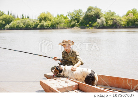 Caring fisherman petting spotted dog, while sitting in boat, fishing. Front view of pensive male hugging shaggy setter, while holding rod in one hand, with bushes on background. Concept of friendship. 107507037