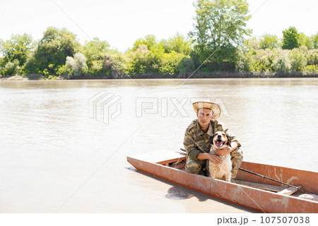 Caring young man in camo posing at camera, while hugging nice spotted setter, while sitting in boat on calm river. Front view of male pet owner petting cute dog, while fishing. Concept of association. 107507038