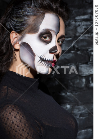 Mysterious dark haired woman with skull makeup looking at camera. Portrait of caucasian girl with scary make up with enigmatic look, in front of dark brick wall. Concept of Halloween. 107507050