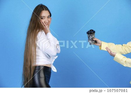 Side view of pretty young woman with very long hair covering face from joy, while unrecognisable male hand in yellow shirt offering free coffee in paper cup, isolated on blue. Concept of emotions. Side view of pretty young woman with very long hair covering face from joy, while unrecognisable male hand in yellow shirt offering free coffee in paper cup, isolated on blue. Concept of emotions. 107507081