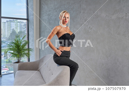 Stunning blonde female in designed strapless top posing, while leaning on sofa edge at home. Side view of pretty woman with hairstyle sitting seductively on couch with hand in pocket. Fashion concept. Stunning blonde female in designed strapless top posing, while leaning on sofa edge at home. Side view of pretty woman with hairstyle sitting seductively on couch with hand in pocket. Fashion concept. 107507970