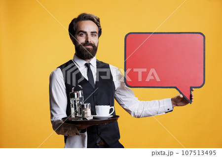 Young employee presents blank speech bubble in studio, working at restaurant and showcasing marketing advertisement with empty billboard copyspace. Waiter shows cardboard icon. Young employee presents blank speech bubble in studio, working at restaurant and showcasing marketing advertisement with empty billboard copyspace. Waiter shows cardboard icon. 107513495