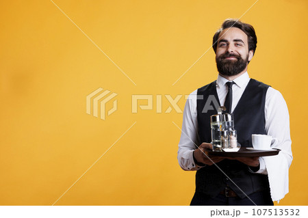 Smiling butler poses with platter in hand, standing against yellow background while he works at five star restaurant. Experienced employee with tray and towel taking orders, serving customers. Smiling butler poses with platter in hand, standing against yellow background while he works at five star restaurant. Experienced employee with tray and towel taking orders, serving customers. 107513532