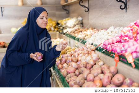 Delighted young Muslim woman purchaser choosing onions in grocery store 107517553