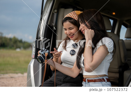 Two pretty Asian female friends are enjoying talking while sitting on a car's open trunk together. Two pretty Asian female friends are enjoying talking while sitting on a car's open trunk together. 107522372