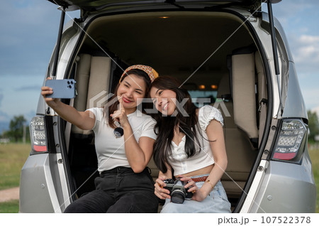 Two joyful Asian women are enjoying their road trip, using a phone to take selfies in an open trunk. 107522378