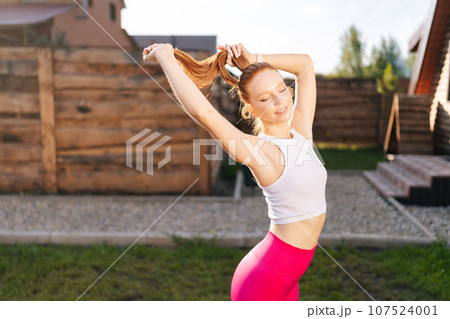 Portrait of beautiful athletic young woman in sportswear living confidently, standing posing with closed eyes on background of green grass on summer sunny day. Concept of wellness, fitness. Portrait of beautiful athletic young woman in sportswear living confidently, standing posing with closed eyes on background of green grass on summer sunny day. Concept of wellness, fitness. 107524001