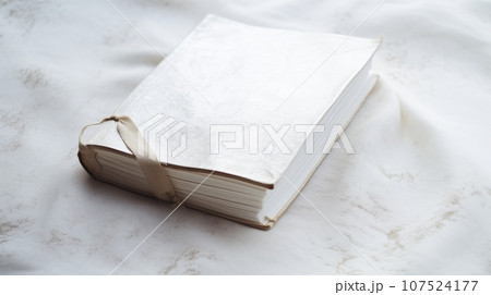 a white book with a ribbon on a white marble surface. The book has a textured cover and the ribbon is tied in a bow. The marble background has gray veins and adds elegance to the image 107524177