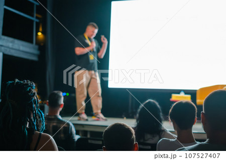 Male speaker standing on stage near big screen and giving presentation to participants sitting in dark hall during business seminar Male speaker standing on stage near big screen and giving presentation to participants sitting in dark hall during business seminar 107527407