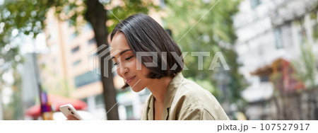 Close up portrait of asian girl sitting in cafe outside, drinking coffee and using smartphone, reading mobile phone 107527917