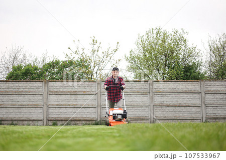 view of man in casual clothes mows lawn with lawn mower at backyard of his house. Husband takes care of garden on spring cloudy day. Modern gasoline garden equipment. Landscaping work view of man in casual clothes mows lawn with lawn mower at backyard of his house. Husband takes care of garden on spring cloudy day. Modern gasoline garden equipment. Landscaping work 107533967