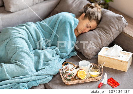 Flu epidemic. A sick woman sleeps under a blanket on the sofa. In front of her there is a tray with a teapot, lemon and honey. Nearby are pills, a thermometer, nasal spray and tissues. Blurry face. Flu epidemic. A sick woman sleeps under a blanket on the sofa. In front of her there is a tray with a teapot, lemon and honey. Nearby are pills, a thermometer, nasal spray and tissues. Blurry face. 107534437