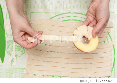 A young woman wraps a strip of puff pastry around an apple circle before baking it in the oven. A young woman wraps a strip of puff pastry around an apple circle before baking it in the oven. 107535871